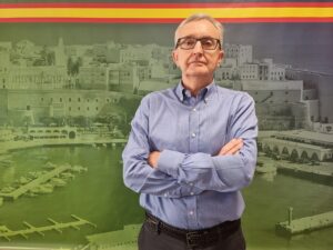 Man wearing glasses and a blue button-down shirt stands with arms crossed in front of a large cityscape mural with a yellow and red stripe at the top of the image.