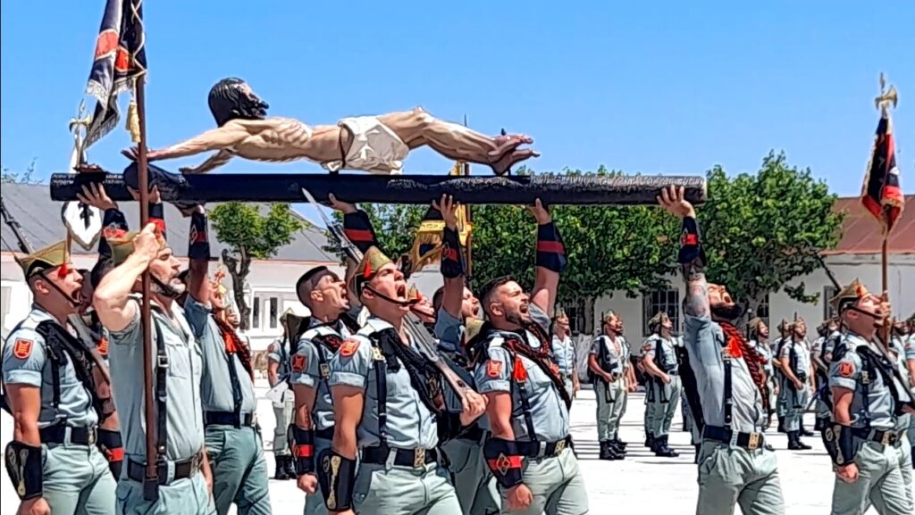 Uniformed soldiers in a parade lift a prone figure on a horizontal beam, imitating a crucifixion pose, flags in the background.