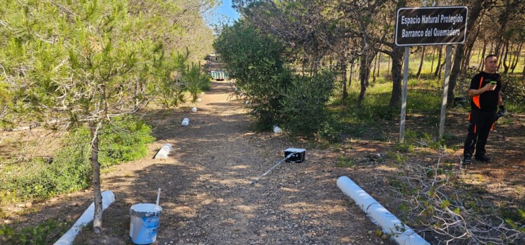 Maintenance worker in orange and black stands beside a gravel path in a protected natural area, with tubes and a toolbox along the trail.