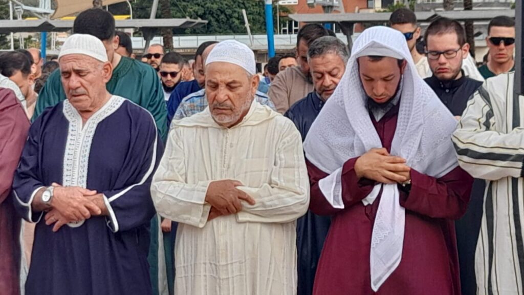 Group of men standing in rows, praying with hands folded over their chests, wearing traditional robes and head coverings outdoors.