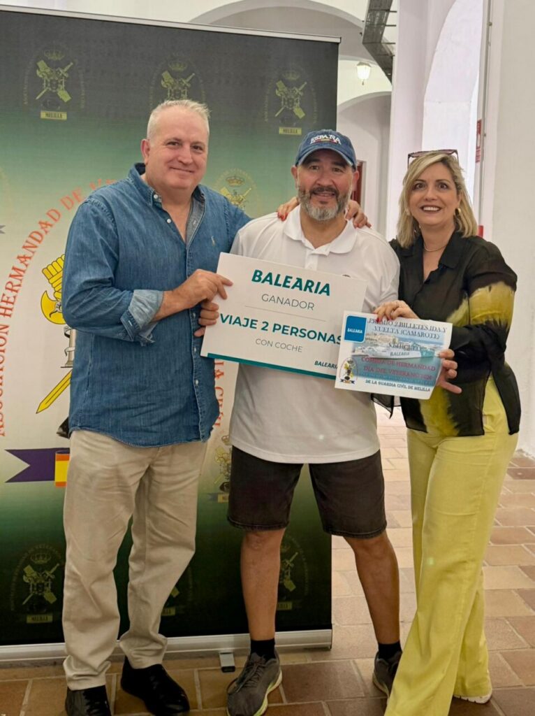 Three people posing with a prize sign: ‘BALEARIA Ganador… Viaje 2 personas’ and a second card, smiling in a bright indoor setting.