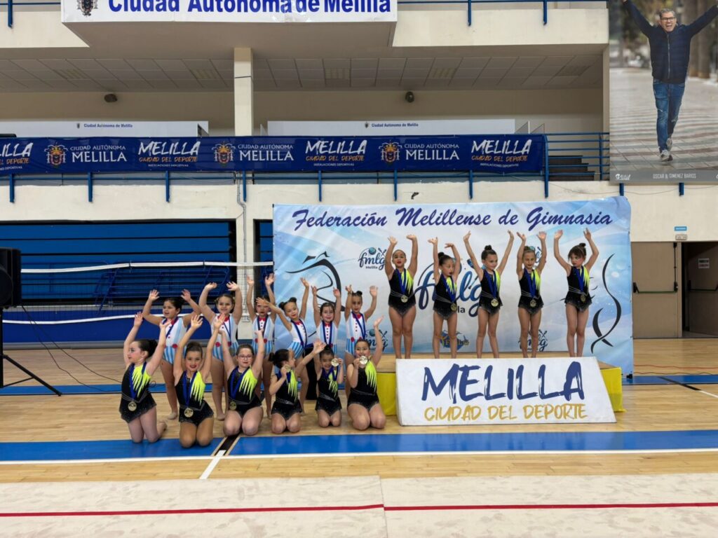 Group of young gymnasts on a podium posing with medals in a gym, banners read Melilla Ciudad del Deporte in the background.