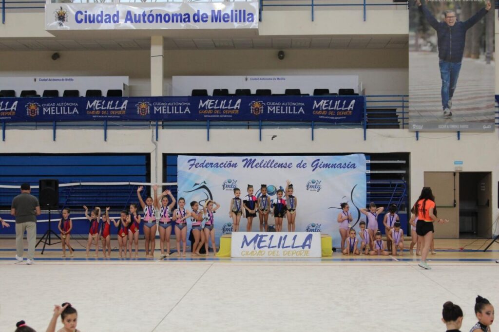 Young gymnasts on a podium posing after a gymnastics event in a sports hall; banner reads Federación Melillense de Gimnasia in the background.