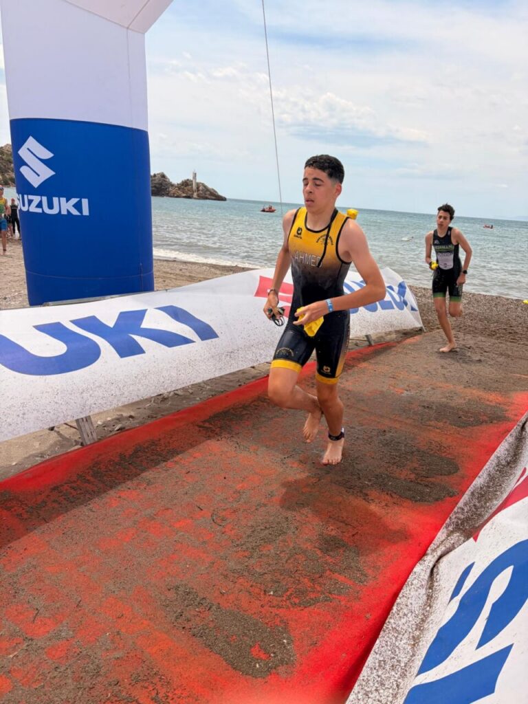 Male athlete crosses the beach finish line on a red carpet under a Suzuki arch, with another competitor behind and the ocean in view.