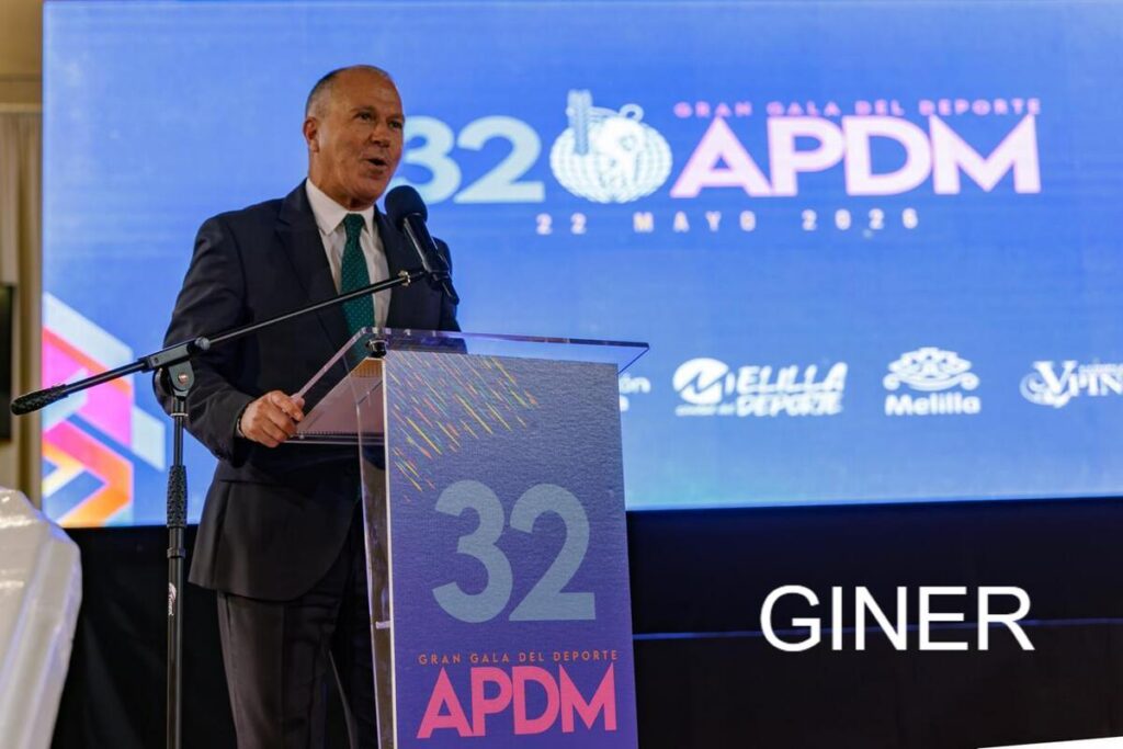 Man in a dark suit speaks at a clear acrylic podium on stage with a blue APDM backdrop showing '32 APDM' and 'Gran Gala del Deporte'.