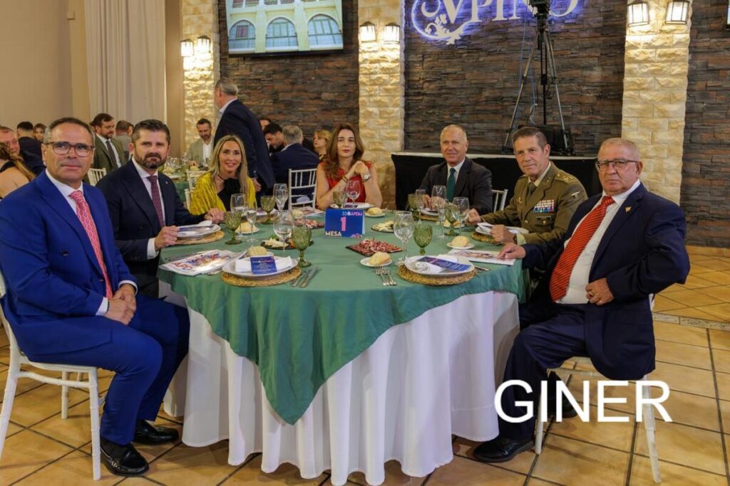 Seven adults in formal attire around a round banquet table; includes a military officer, glassware and appetizers visible.