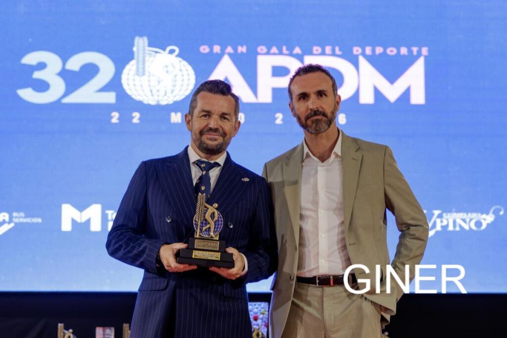 Two men in suits pose on stage after receiving a trophy at a sports awards gala, blue backdrop with event logo on display.