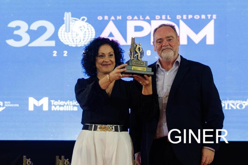Two presenters on a stage hold up a trophy together during a sports awards ceremony, smiling, with a blue backdrop and event logos behind them.