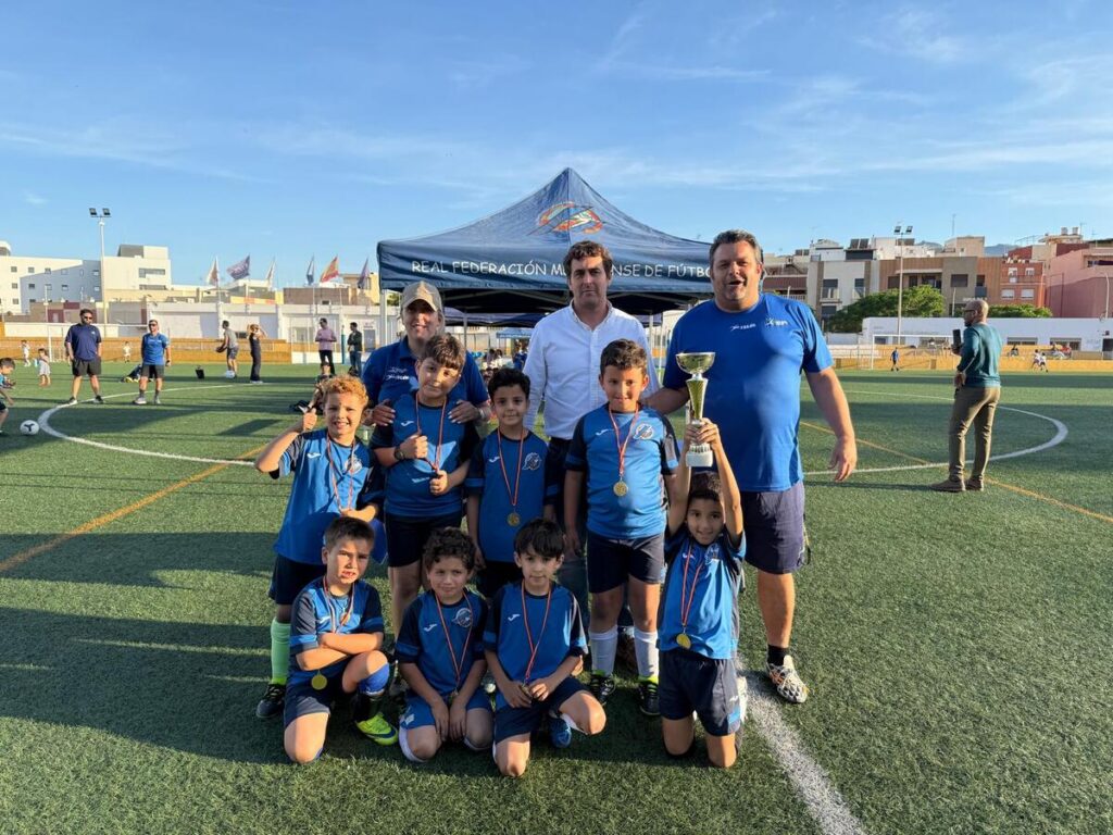 Group of young soccer players in blue uniforms with medals, posing with coaches on a sunny turf field after a game; one child holds a trophy high.