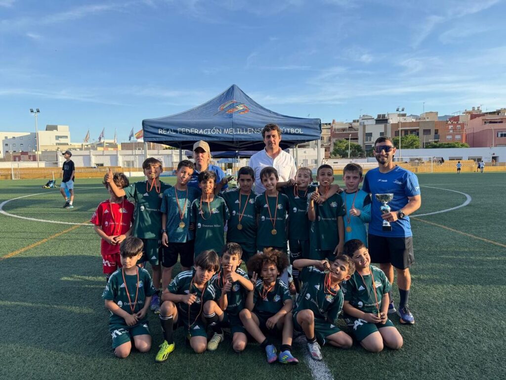 Youth soccer team in green uniforms posing with medals on a turf field, coaches nearby under a blue tent that reads Real Federación Melillense Fútbol; trophy held by a coach on the right.