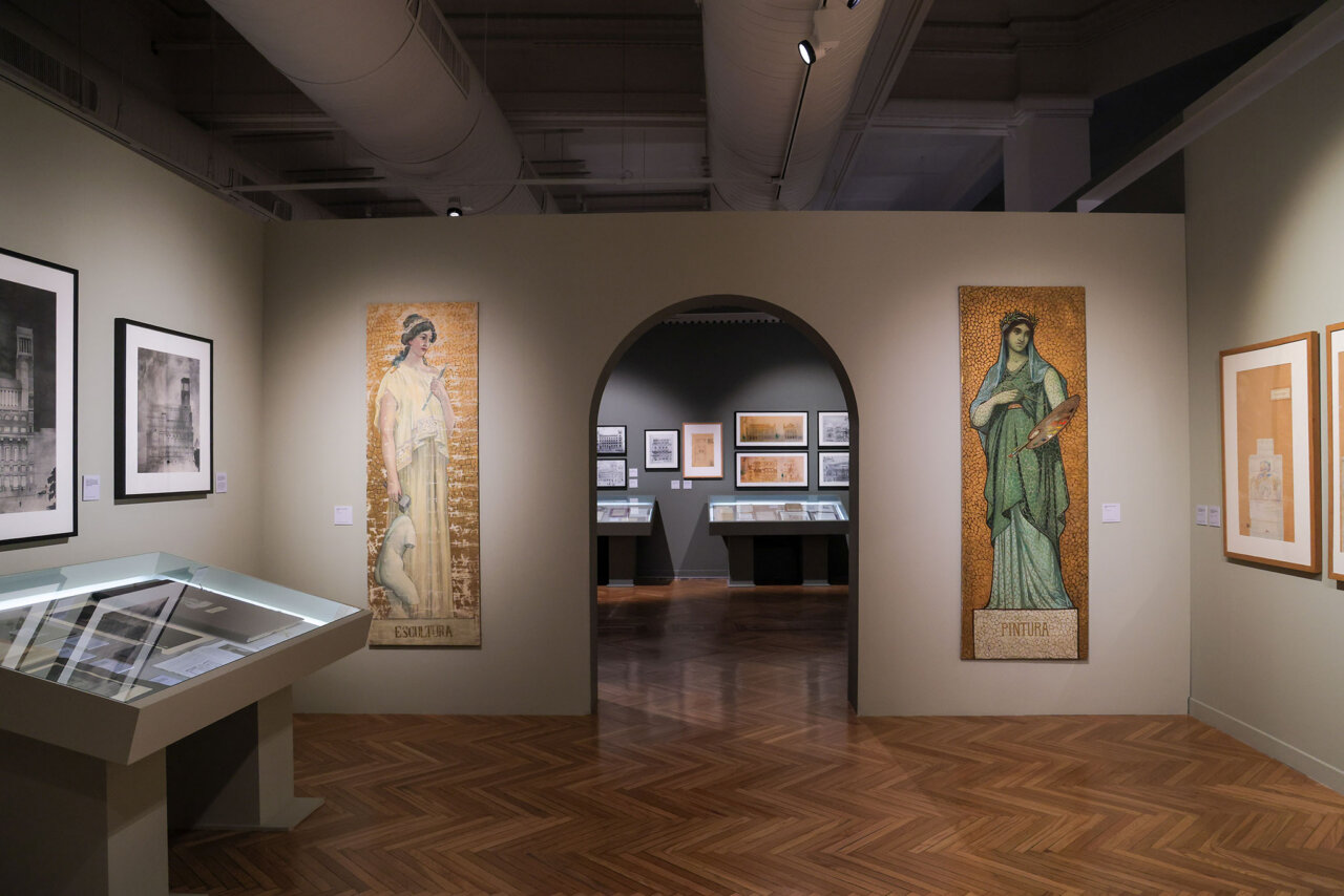 Museum gallery with an arched doorway opening to a side room, flanked by tall mosaic panels of a woman and a saint, and glass display cases along the walls.