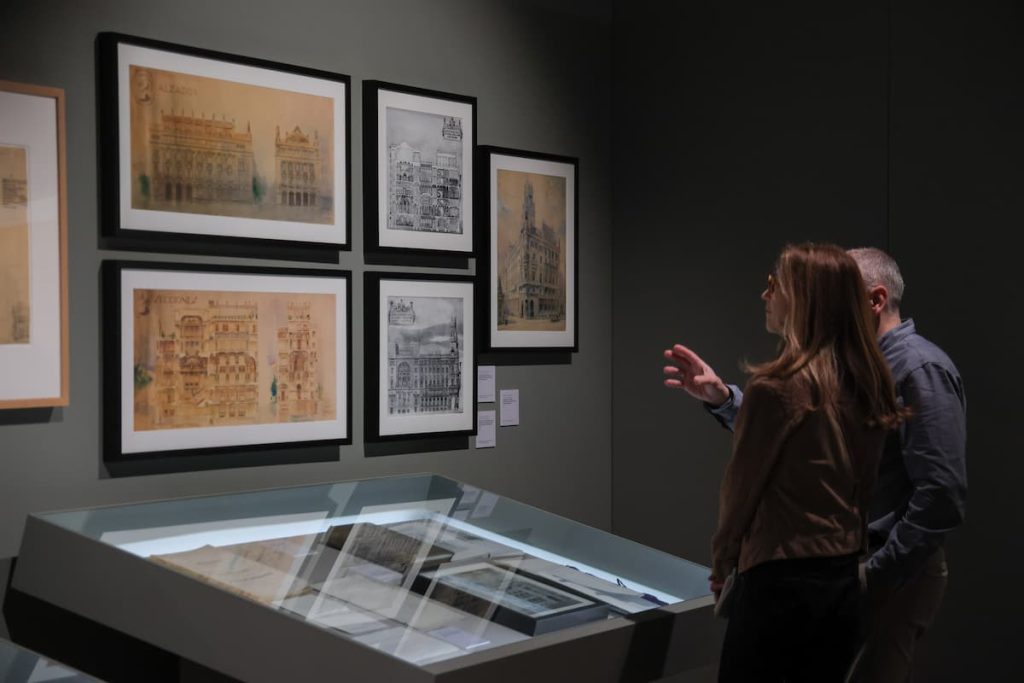 Two visitors in a gallery examine framed architectural prints on a dark wall, with a glass display case in the foreground.
