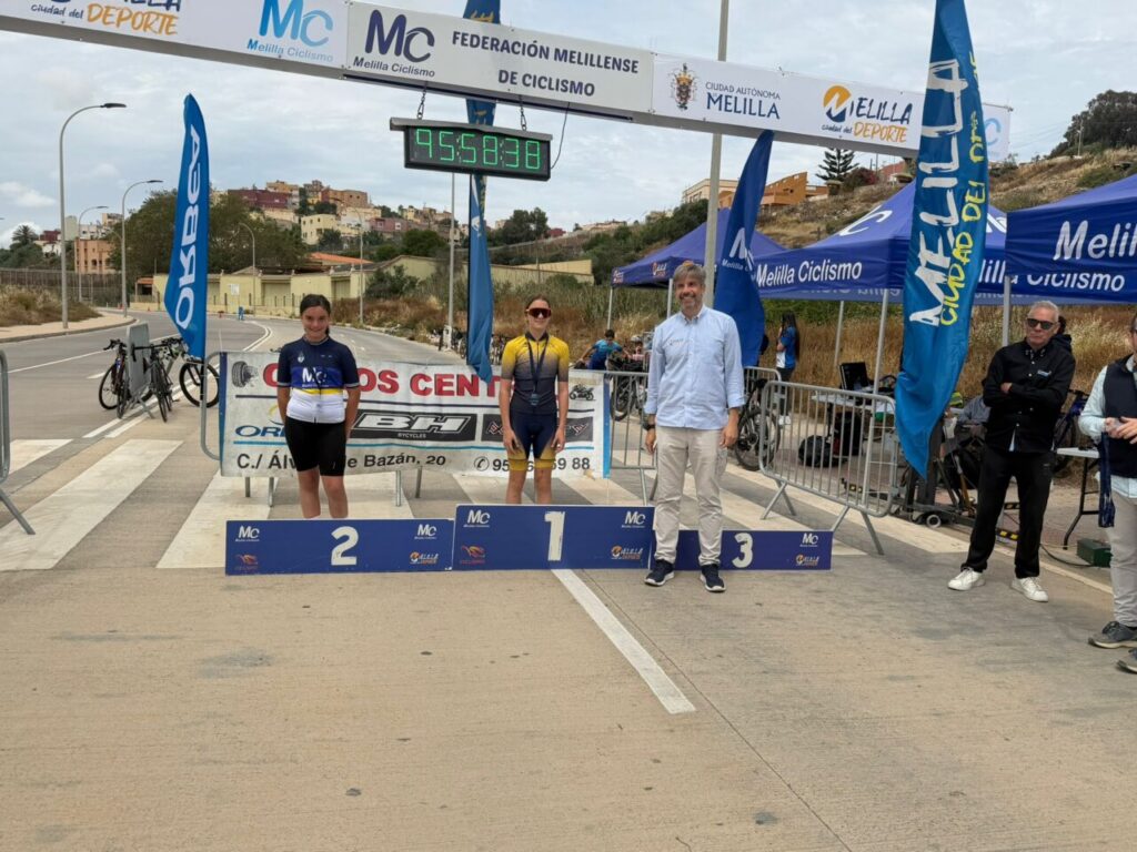 Podium at a cycling race finish: two female cyclists (2nd left, 1st center) with a man standing beside the 3rd place step under the finish banner.