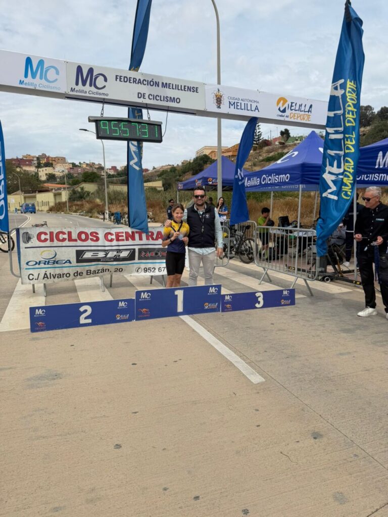 Finish-line podium for a cycling race with 1st place center and 2nd/3rd spots to the sides; two people pose for a photo under event banners.