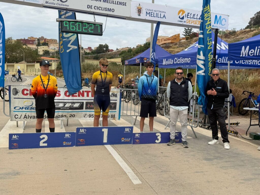 Three young cyclists stand on a finish-line podium with positions 1, 2, and 3, medals around their necks, under Melilla Ciclismo banners and a digital timer reading 96:02:27.
