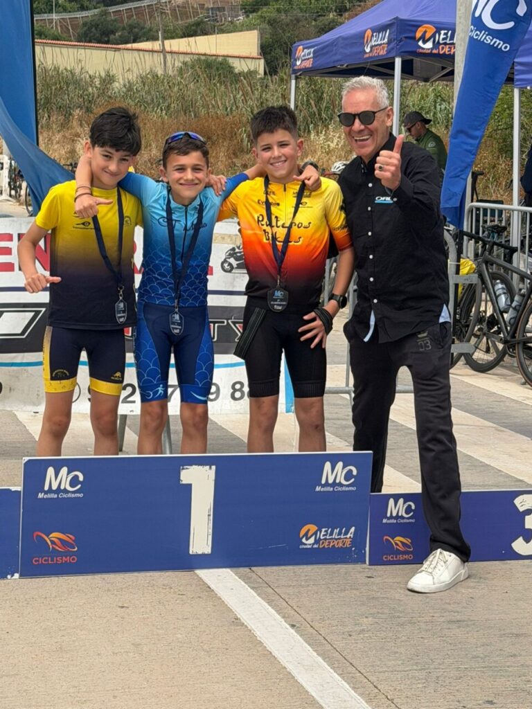 Three young cyclists on a winners' podium with medals, posing with an older man giving a thumbs up; event banners and racing team logos in the background.