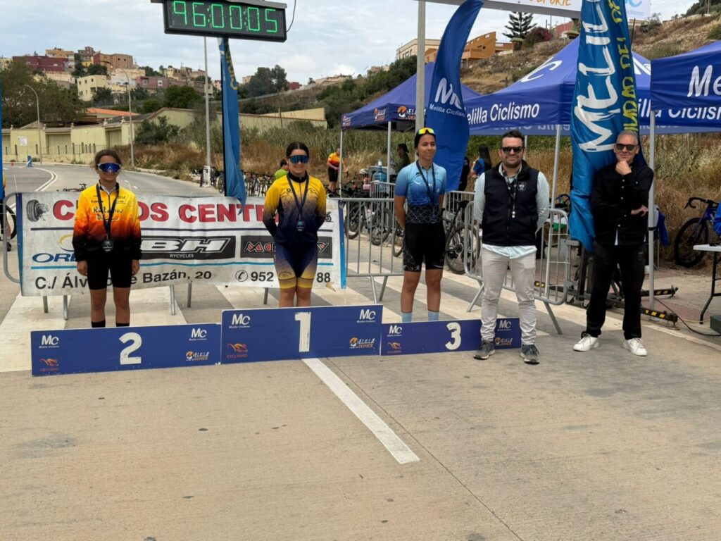 Podium finishers pose with their medals: 1st, 2nd, and 3rd place, flanked by organizers under blue banners at a cycling event.