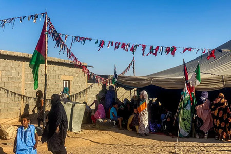 Dusty refugee camp scene with a large canopy shelter, colorful bunting, and groups of people sitting on the ground near stone buildings.