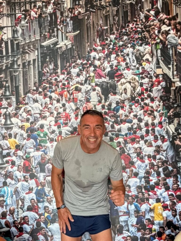 Smiling man in a gray T-shirt and navy shorts poses in front of a wall-sized photo of a crowded street filled with runners wearing white outfits and red sashes.