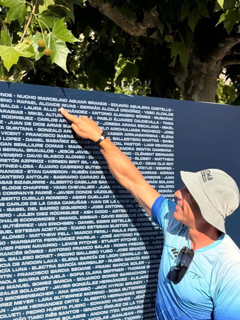 Man in a blue shirt and hat points at a large blue wall covered with white names outdoors beneath a leafy tree.