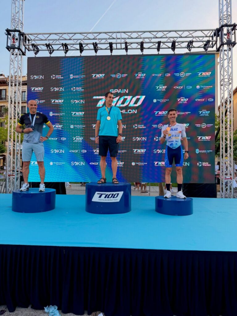 Three male medalists stand on blue podiums during a triathlon awards ceremony on a bright stage with a large digital banner behind them reading 7100 Triathlon World Tour Spain.