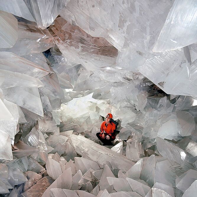 Person wearing an orange helmet and jacket sits among large clear crystal formations in a cave.