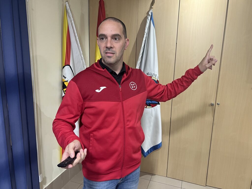 Man in a red athletic jacket gestures to the right while holding a remote, standing in a room with flags in the background.