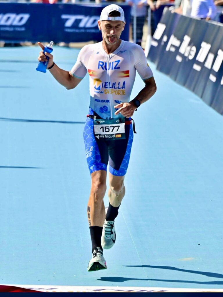 Triathlete Ruiz finishes a race on a bright blue track, holding a water bottle and wearing a Spain-themed jersey with bib 1577 (Luis Miguel).