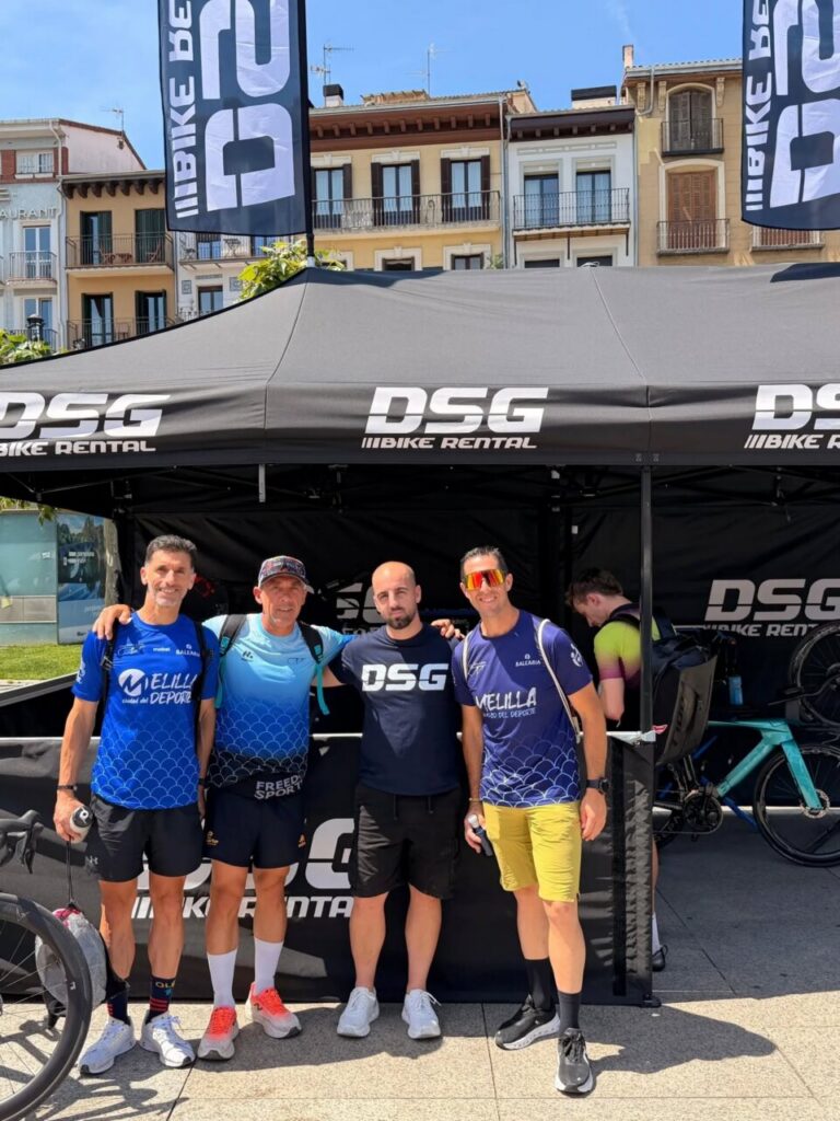 Five male cyclists pose together under a DS G bike rental tent at an outdoor event, with buildings in the background.