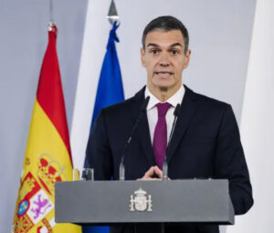 Man in a dark suit and burgundy tie delivers a speech at a podium with a crest, flanked by the Spanish flag and a blue flag behind him.