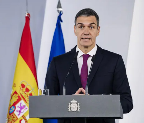 Man in a dark suit and burgundy tie delivers a speech at a podium with a crest, flanked by the Spanish flag and a blue flag behind him.
