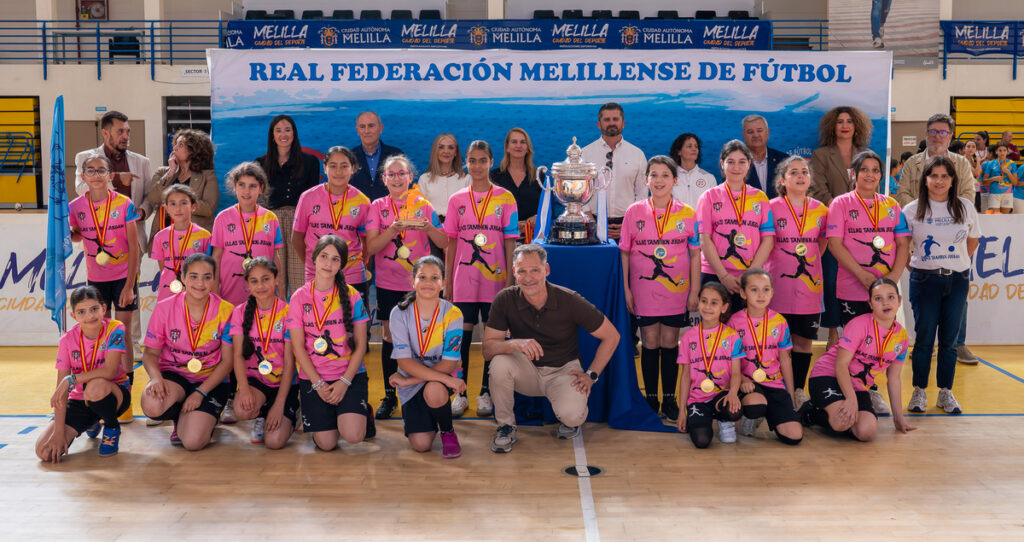 Girls in pink shirts with medals pose around a large trophy on a gym floor during a football award ceremony, with a banner in the background reading 'Real Federación Melillense de Fútbol'.