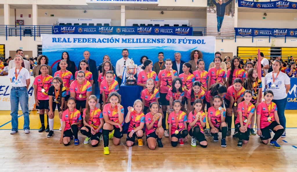 Group of young girls in pink football jerseys with medals, posing in a gym with a large trophy and a banner reading Real Federación Melillense de Fútbol.
