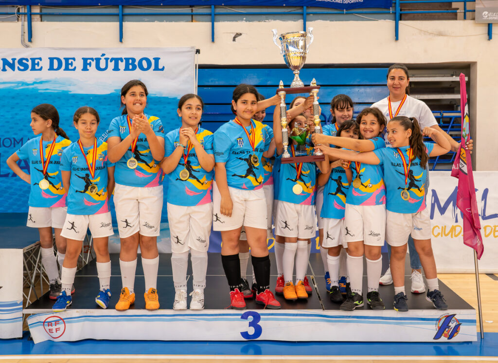 Youth girls' soccer team on a podium, blue jerseys and medals, lifting a large trophy as a coach looks on in the background.