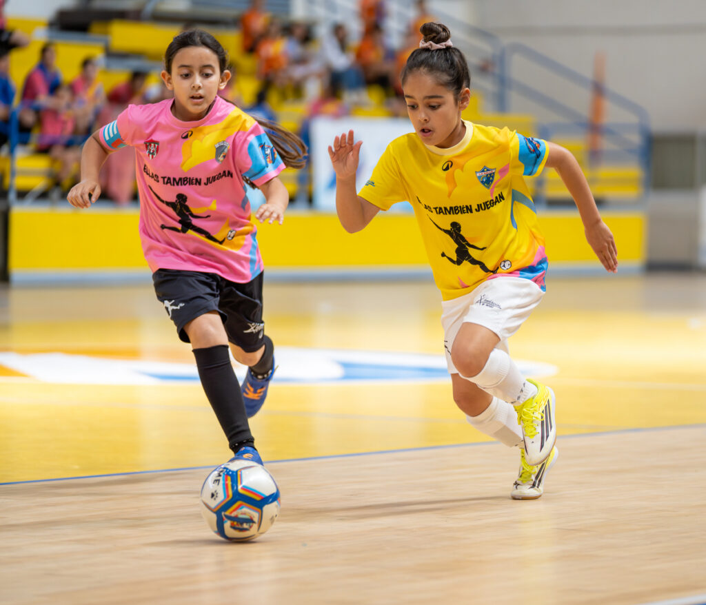Two young girls play indoor soccer, one in a pink jersey and black shorts dribbling the ball while a teammate in yellow races alongside on a bright gym floor.