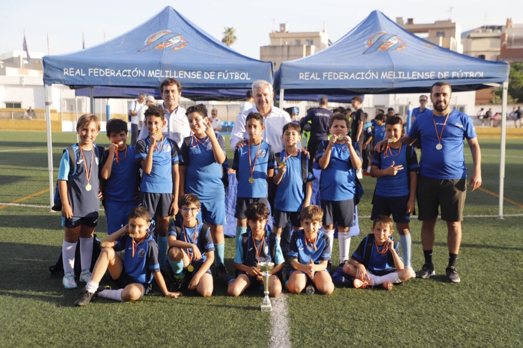 Youth soccer team of children posing with medals and a trophy under blue tents at a field event.