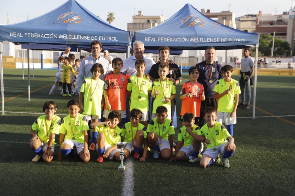 Youth soccer team with medals and a trophy, posing on a green pitch under blue event tents with adults behind them.