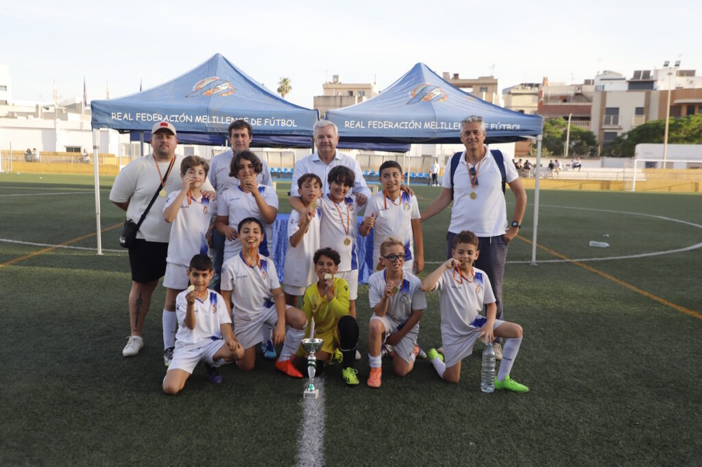 Group of young soccer players with coaches posing on a field after winning, medals around necks and a trophy in front.