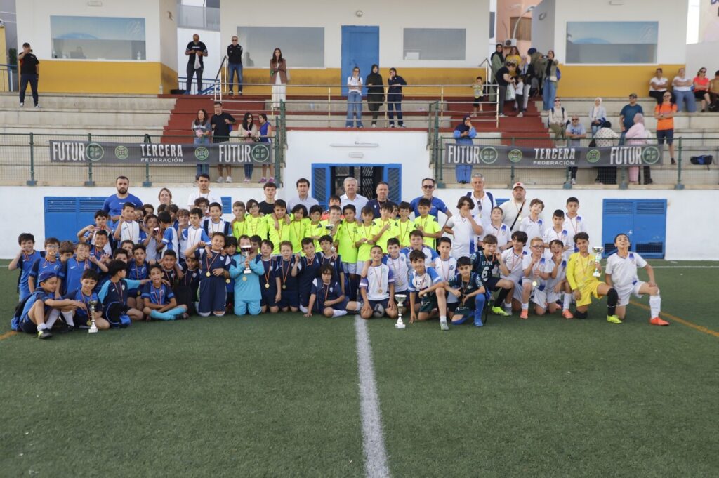Group of young soccer players and coaches pose with trophies on a turf field, spectators in bleachers behind them.