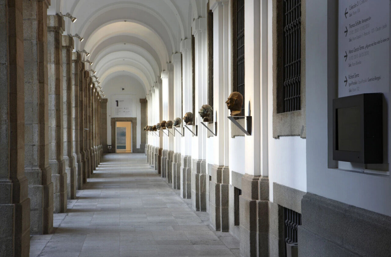 Long arcade at Museo Reina Sofía with bust sculptures on shelves along white columns and arches in a well-lit corridor.
