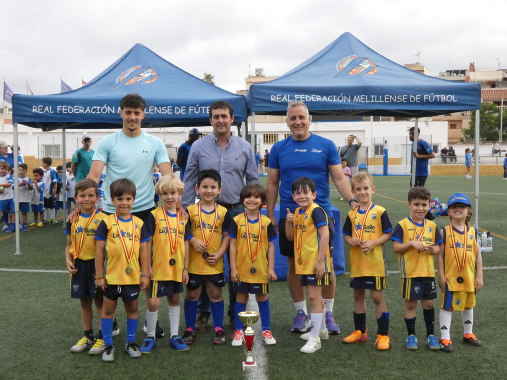 Young soccer team in yellow jerseys with medals posing with coaches on a grassy field; a trophy sits at their feet with blue tournament tents in the background.