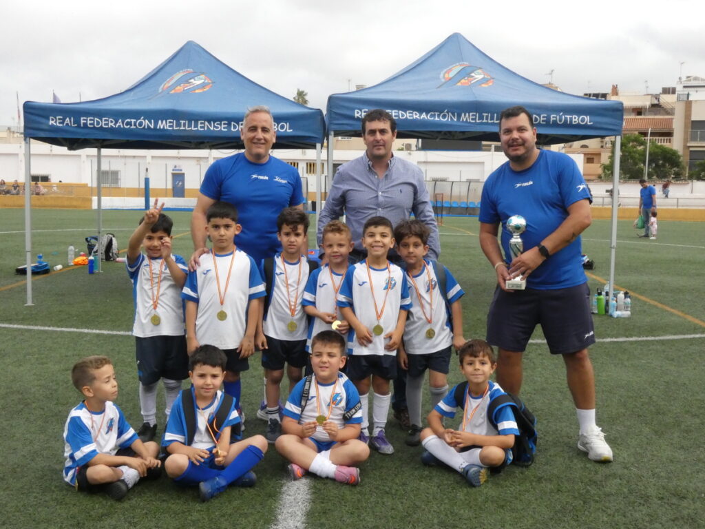 Kids soccer team with medals posing for a photo on a turf field, coaches behind, tents reading Real Federación Melillense de Fútbol.