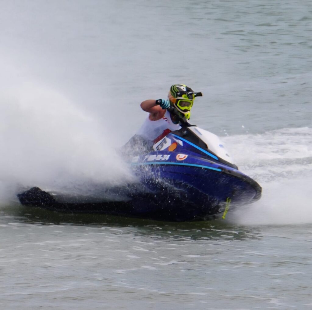 Jet ski rider in a neon helmet leans into a turn on a blue jet ski, spraying water behind them on a lake.