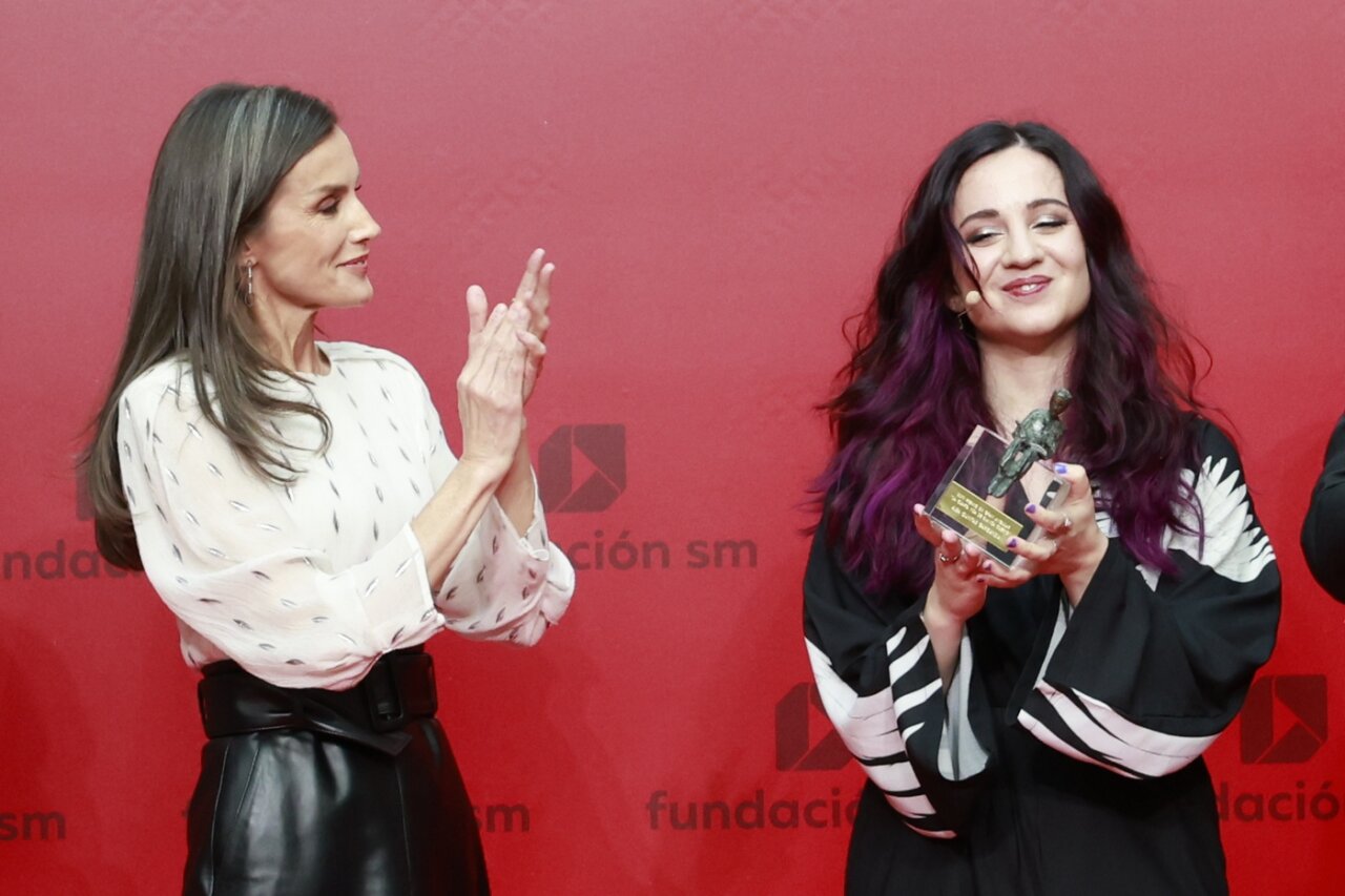 Two women at an award ceremony on a red backdrop; one claps while the other holds a trophy and smiles, celebrating a win.