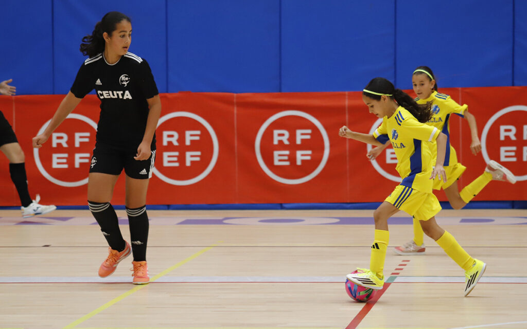 Young girl in yellow dribbles a ball on an indoor court, chased by a girl in black with a Ceuta jersey, red banners in the background.