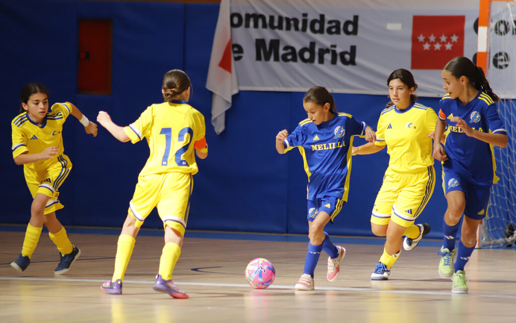 Girls' futsal match: players in yellow and blue vie for the pink ball on an indoor court, with a 'Comunidad de Madrid' banner in the background.