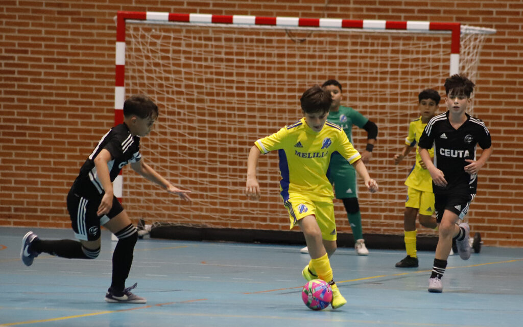 Indoor futsal game: a boy in yellow dribbles a bright pink ball toward the goal, chased by opponents in black and yellow kits.