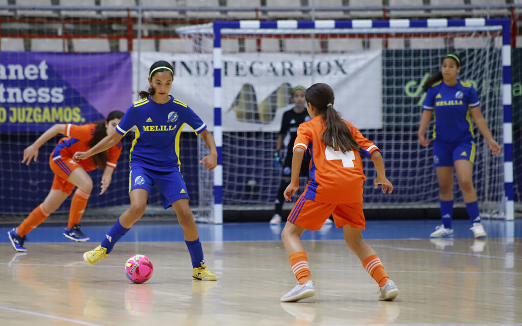 Young female futsal players in blue and orange kits chase a bright pink ball on an indoor court, with a goal and net in the background.