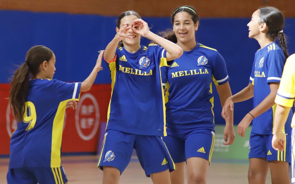 Female youth team in blue and yellow uniforms celebrate a goal indoors, with MELILLA on their jerseys.
