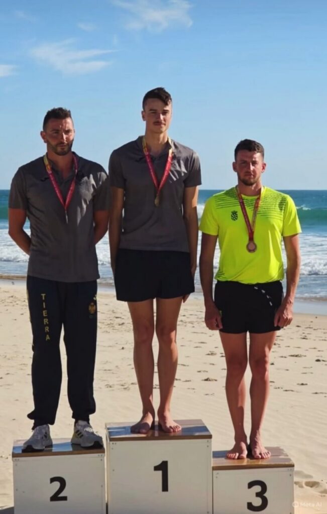 Three male athletes stand on a beach podium: 1st place center, 2nd place left, 3rd place right, each with medals, ocean and sand in the background.